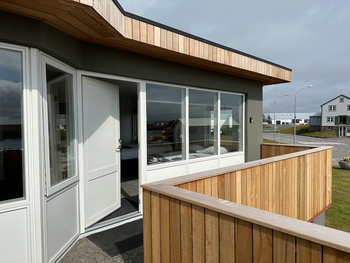 A spacious balcony is accessed through large glass doors, offering views of the surrounding area. The modern exterior features natural wood siding, complemented by a light-colored façade. The entrance area is clean and welcoming, with flooring that transitions smoothly to the outdoors.