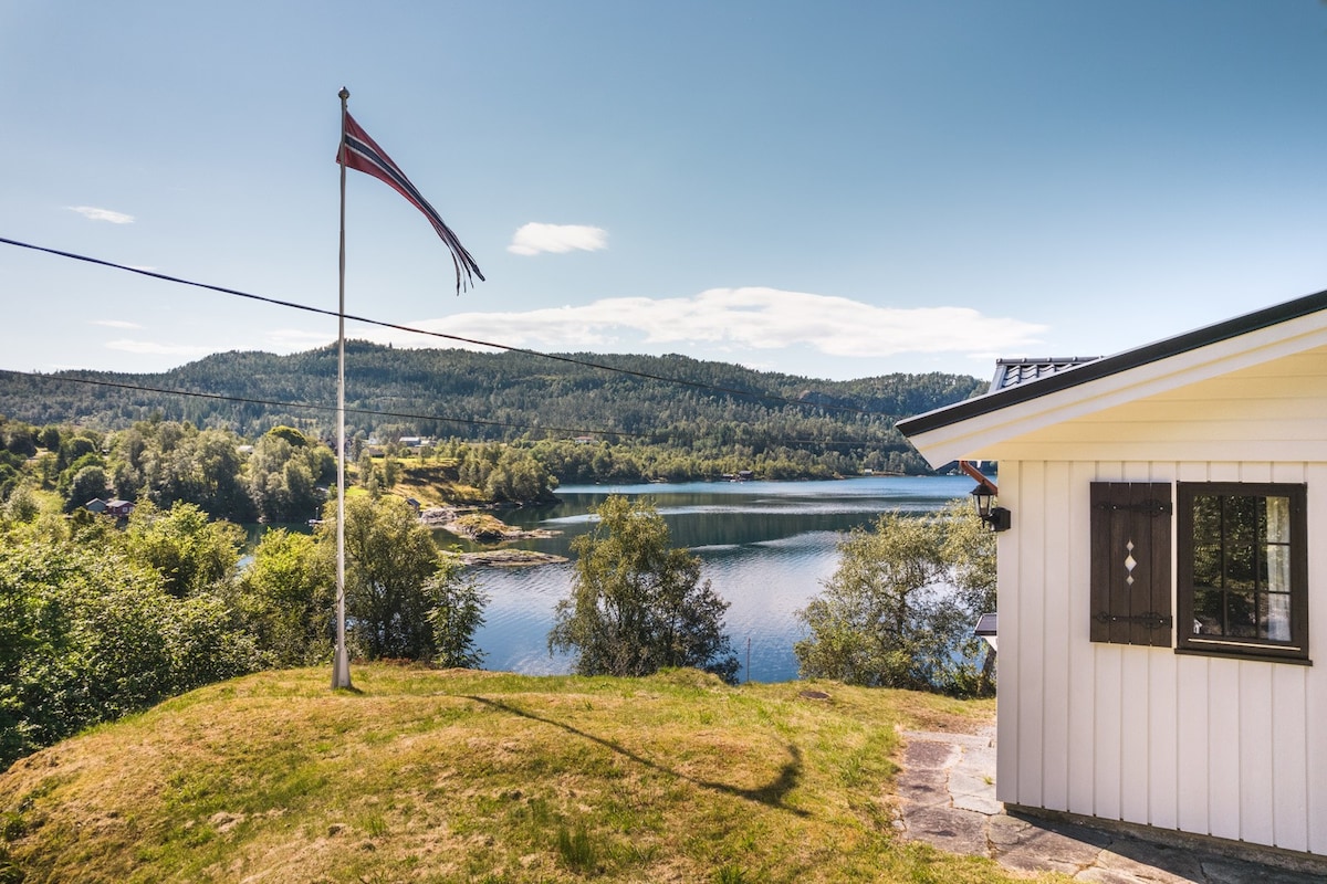 A serene view of Hindenesfjorden is displayed, with gentle hills and trees surrounding the water. A Norwegian flag waves in the foreground, complemented by a white cabin featuring a small window. Clear skies and soft sunlight enhance the tranquil landscape.