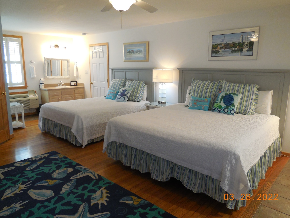 A spacious room features two beds, each with crisp white bedding and decorative pillows. A lighted vanity area with a mirror is visible in the background. A colorful rug with ocean-themed designs lies on the floor, enhancing the room's coastal charm.