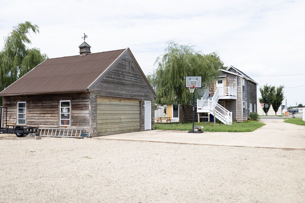 A rustic farmhouse is viewed alongside an adjacent two-story building. A basketball hoop is situated on the paved driveway. The exterior features wood siding with a gabled roof and green grass lining the area. Ample parking space is available in front of the structures.