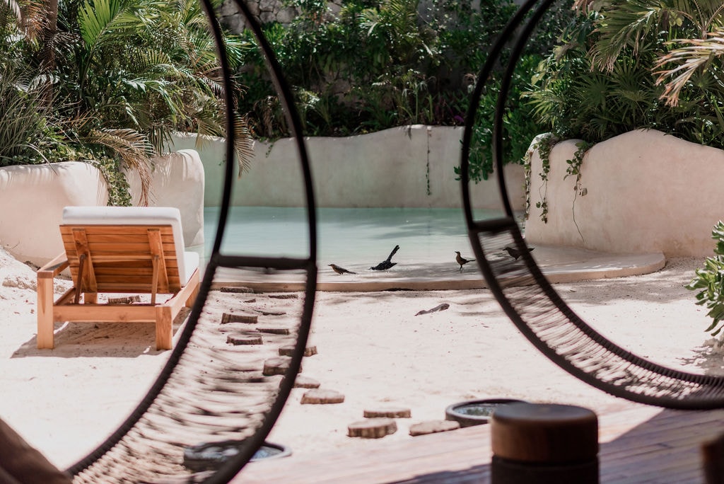 A tranquil private pool is framed by lush tropical plants, creating a serene atmosphere. A wooden lounge chair is positioned nearby on sandy ground, inviting relaxation. The soft curves of the pool and surrounding landscape blend harmoniously, emphasizing a peaceful retreat.