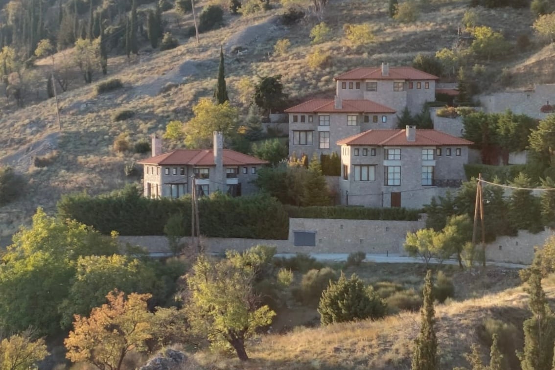 A scenic view of multiple rustic villas nestled among hills, featuring terracotta roofs and large windows. Lush greenery and trees surround the area, enhancing the natural landscape. The property is set against a backdrop of soft, rolling hills under a clear sky.