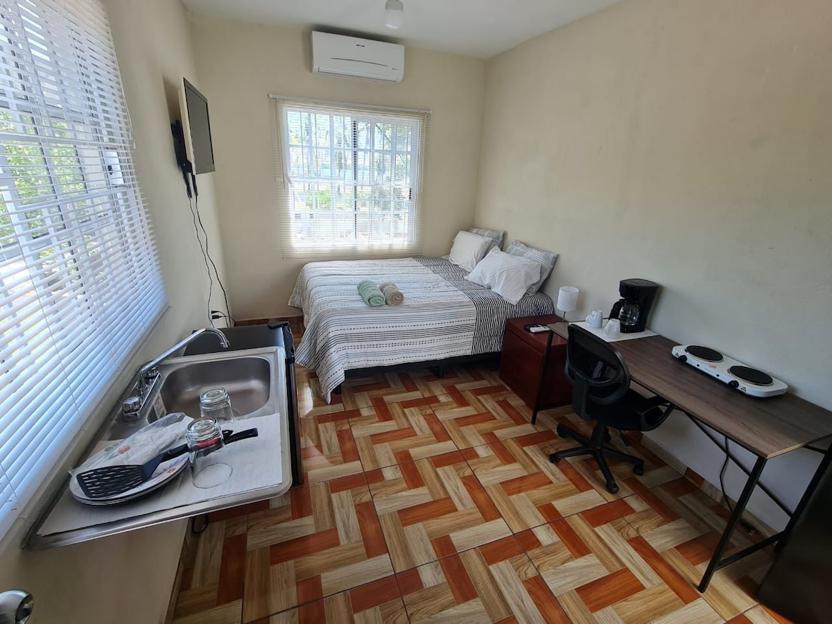 A well-lit room features a spacious bed covered in neutral linens, accompanied by two towels neatly placed on top. A small kitchenette with a sink is in the corner, while a desk with a chair and a coffee maker provides convenient amenities. Large windows offer natural light.