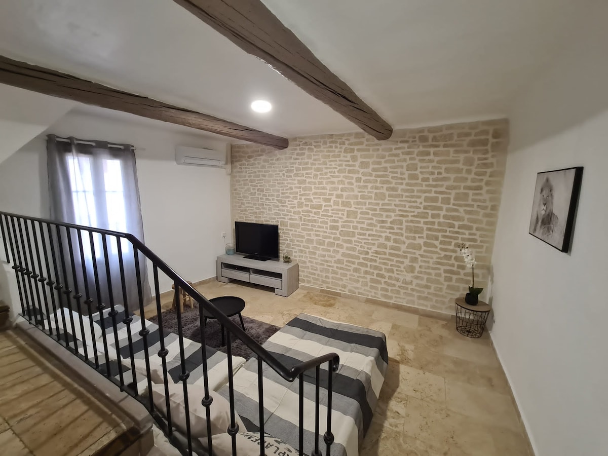 A spacious living area features stone walls and wooden beams. A modern television is positioned on a low cabinet, while a simple black table is placed nearby. Natural light enters through a window covered with sheer curtains, creating a welcoming environment.
