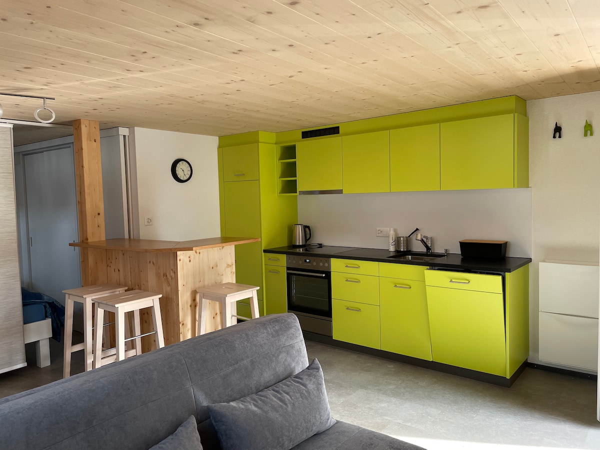 A modern kitchen showcases vibrant green cabinets and sleek black countertops. Essential appliances, including a kettle and coffee maker, are arranged along the counter. A wooden bar with high stools provides additional seating, while a clock indicates the time on the wall.