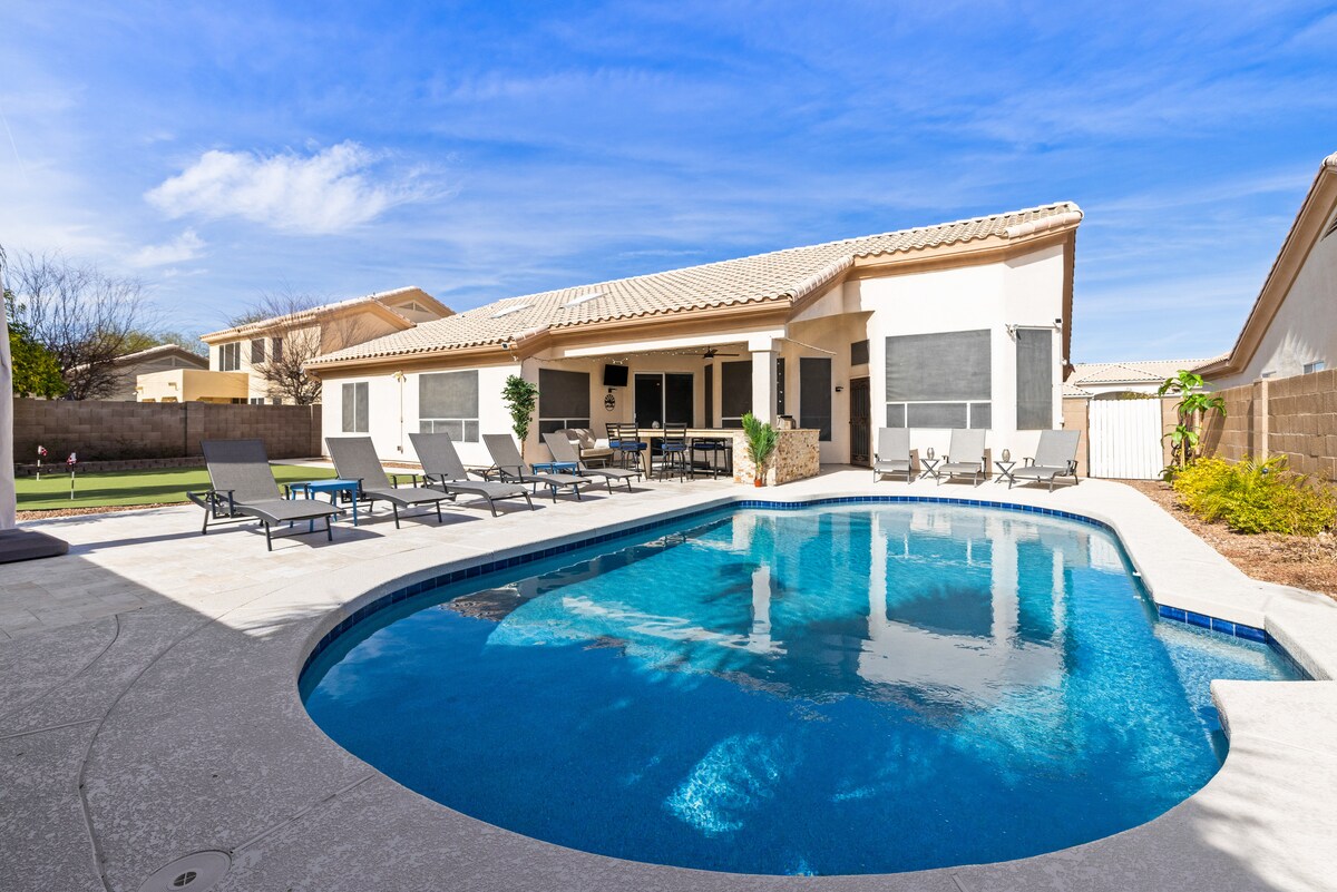The outdoor space showcases a newly remodeled heated saltwater pool, surrounded by multiple lounge chairs. A spacious patio area is visible, featuring seating and a dining space, with the house providing a serene backdrop framed by clear blue skies.