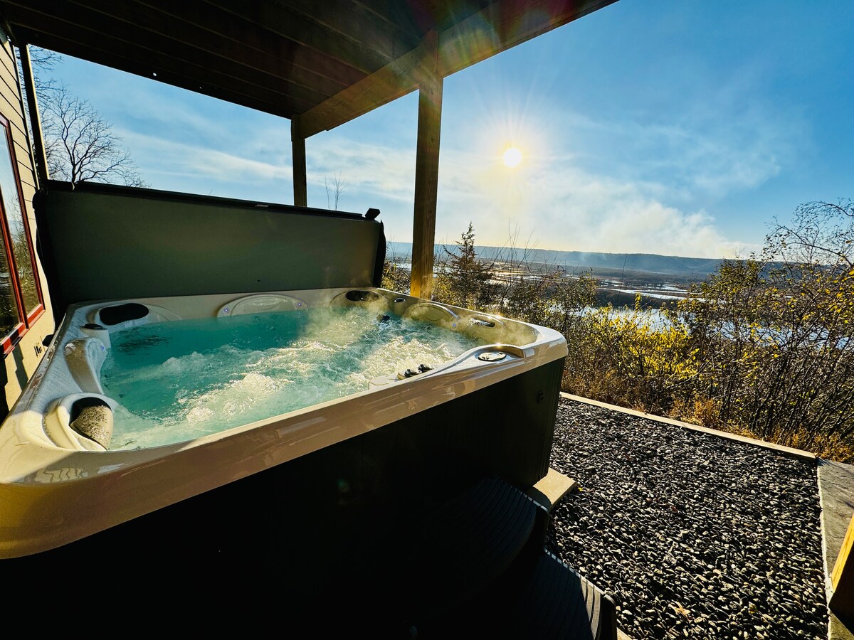 A private outdoor hot tub is shown on a deck, with steam rising and water bubbling in the center. The surrounding area features natural stone landscaping, and a panoramic view of the river is visible in the background, illuminated by the setting sun.