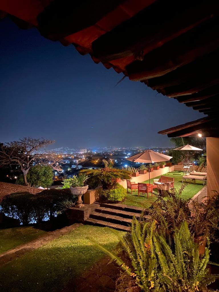A nighttime view reveals a garden area featuring well-maintained grass, stone steps leading to seating arrangements under umbrellas, and potted plants. The twinkling city lights are visible in the background, creating a serene atmosphere enhanced by surrounding greenery.