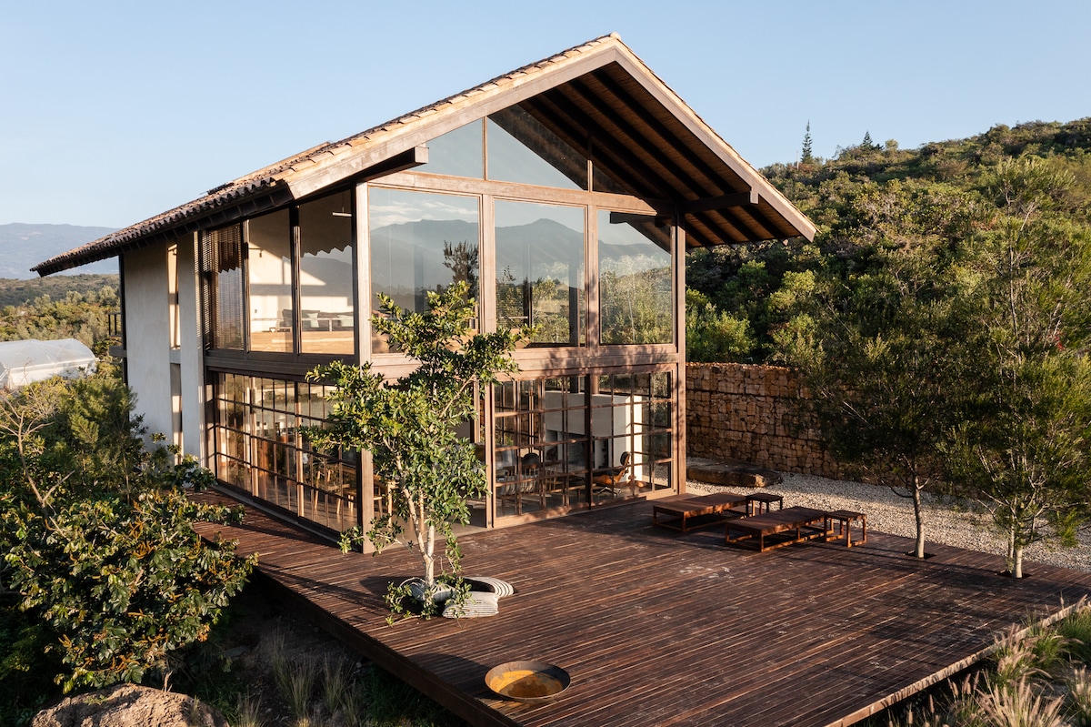 A modern house with large glass windows is set against a backdrop of greenery and distant mountains. A spacious wooden deck extends from the building, furnished with a low table and seating arrangements, inviting outdoor relaxation.