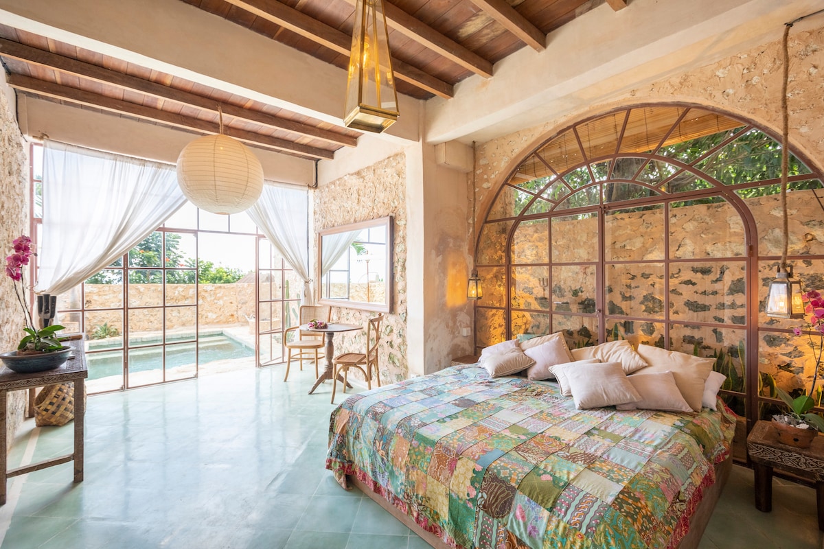 A spacious bedroom features a large bed adorned with a colorful patchwork quilt. Natural light filters through sheer drapes, illuminating the room. An elegant glass door leads to a private pool area, while decorative elements like a woven chair and an orchid accent add charm.