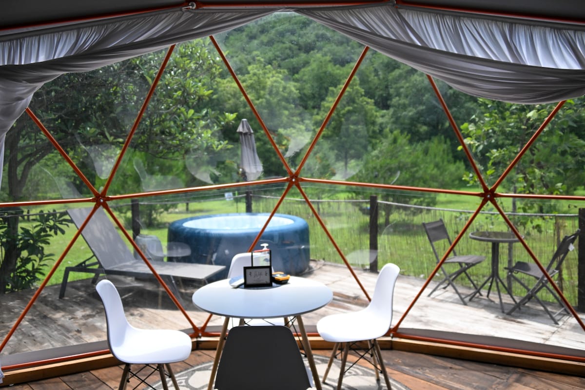 A geodesic dome interior is viewed through large, triangular glass panels, offering a scenic landscape. A circular table with two white chairs occupies the center. Outside, a private hot tub and lounge chairs are visible on a wooden deck, surrounded by lush greenery.