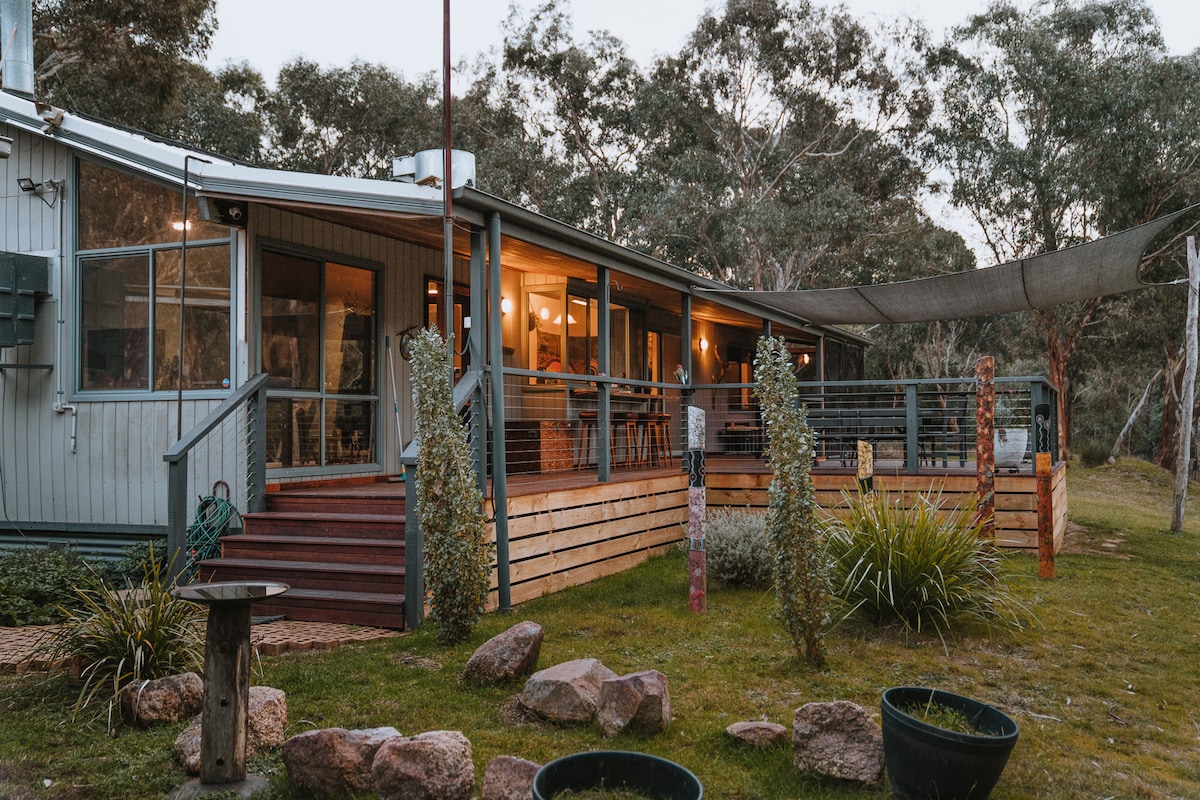 The exterior of the cabin is set amidst native bushland, showcasing a wooden deck with seating. Natural stone and greenery surround the yard, while large windows invite views of the interior. A shade cloth extends over part of the deck, providing a comfortable outdoor space.
