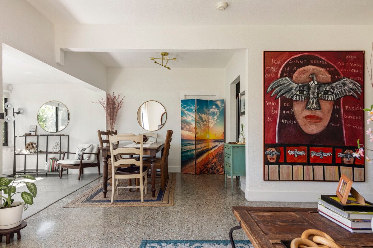 A communal dining area features a long wooden table surrounded by multiple chairs, complemented by decorative wall art. Two round mirrors reflect the space, while a colorful painting depicting a beach scene adds a vibrant touch to the room.