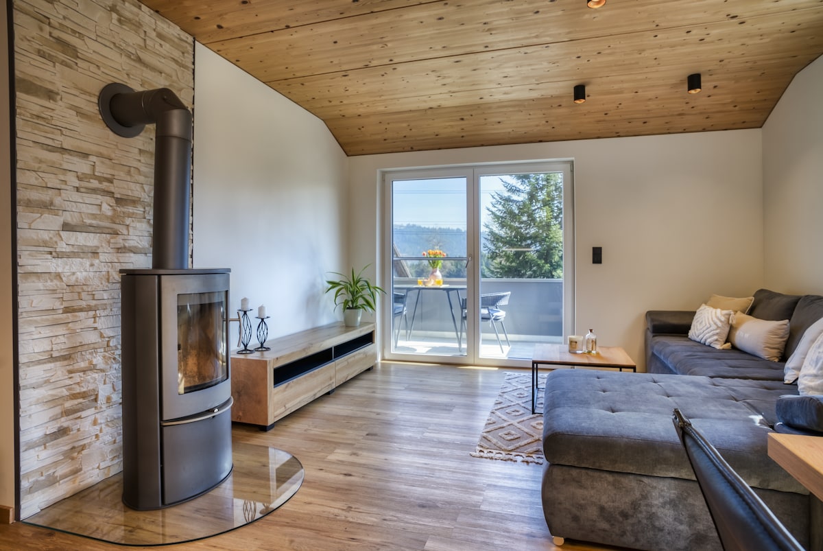 A modern living area is highlighted by a stylish sofa and a stone fireplace, providing a comfortable space for relaxation. Large glass doors open to a balcony, allowing natural light to fill the room and featuring a view of the surrounding landscape.