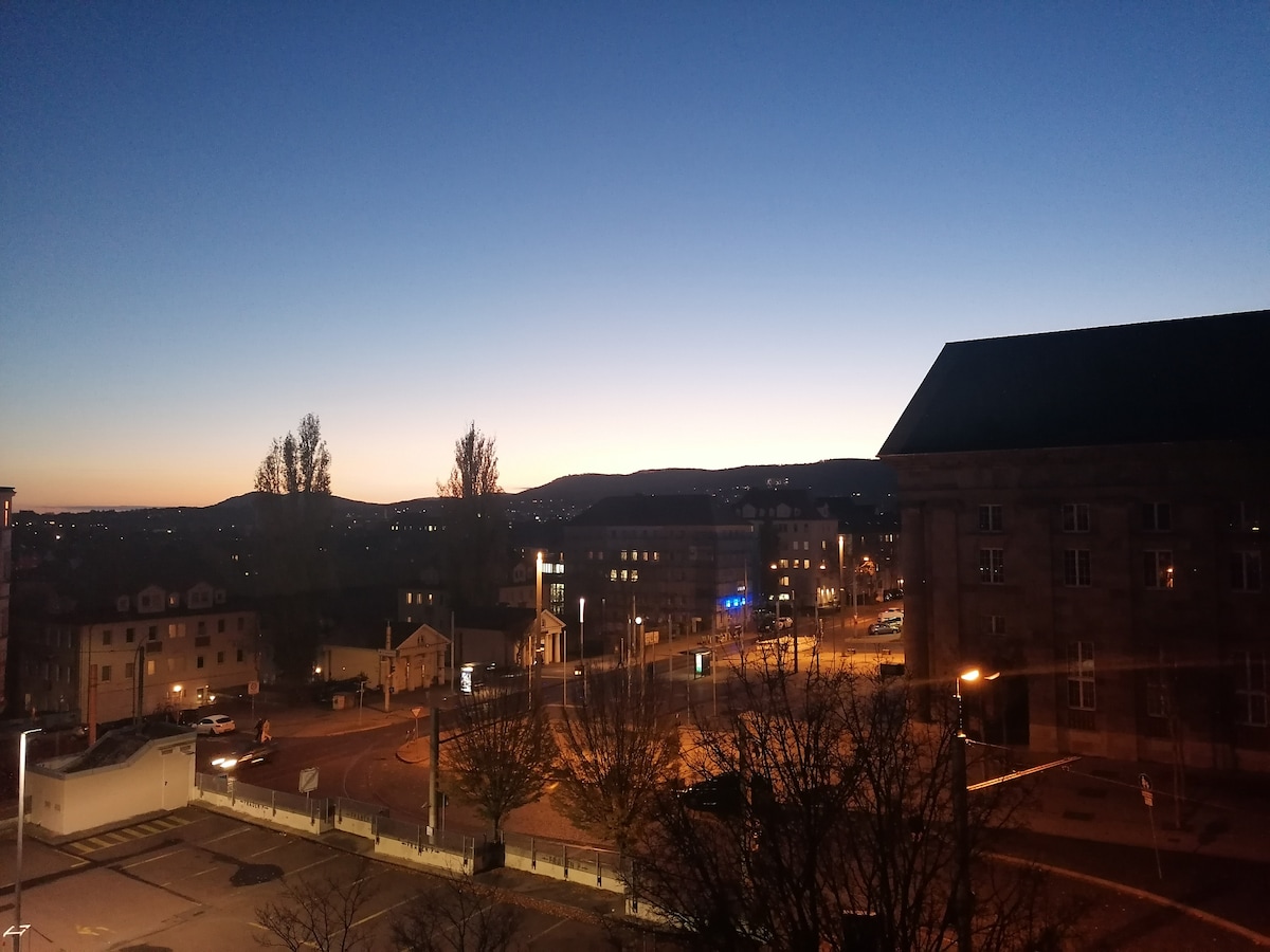 A city skyline is observed as twilight descends, with distant mountains outlined against a fading blue sky. Streetlights illuminate the nearby buildings and roads, creating a calm evening atmosphere.