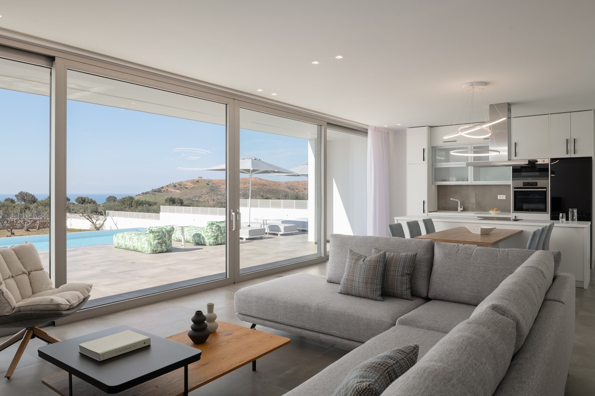 A contemporary living area is showcased, featuring large glass doors that open to a scenic view of the outdoors. Comfortable grey sofas are arranged around a wooden coffee table, with a light fixture overhead. A modern kitchen is visible in the background, integrating seamlessly with the living space.