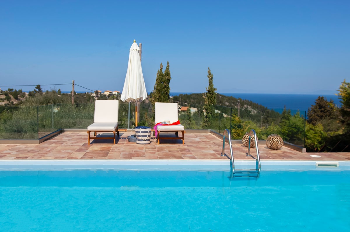 A private outdoor area features a sparkling swimming pool with clear blue water. Two lounge chairs are positioned beside the pool, accompanied by a sun umbrella. A distant view of the sea is visible beyond the landscaped surroundings.