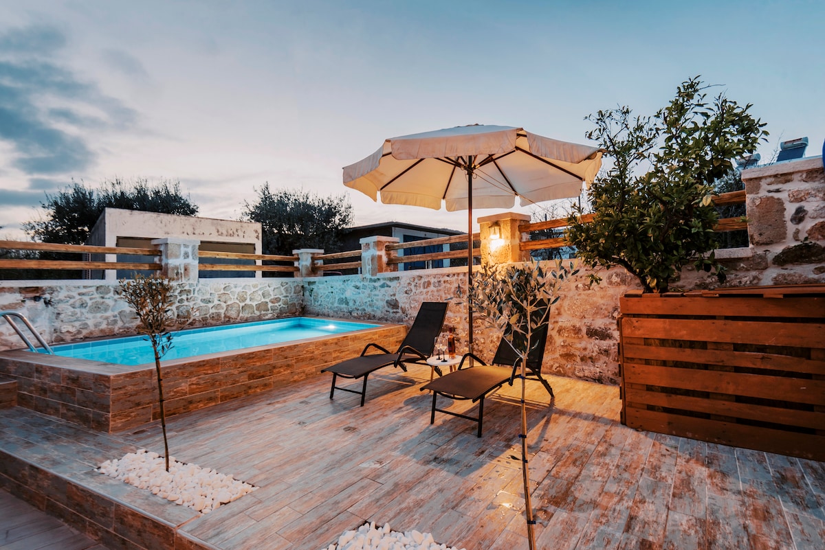 A private swimming pool is framed by natural stone walls, featuring two lounge chairs positioned under an umbrella. Soft lighting illuminates the area as greenery enhances the tranquil outdoor setting.