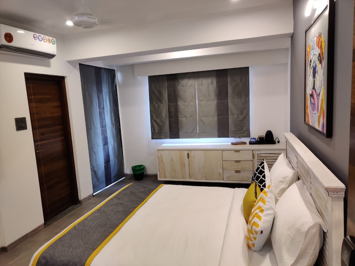 A modern bedroom features a spacious bed adorned with white linens and a yellow accent pillow. A wooden sideboard runs along one wall, while dark curtains partially cover a window. Air conditioning is visible above the bed, and a basket is placed on the floor.