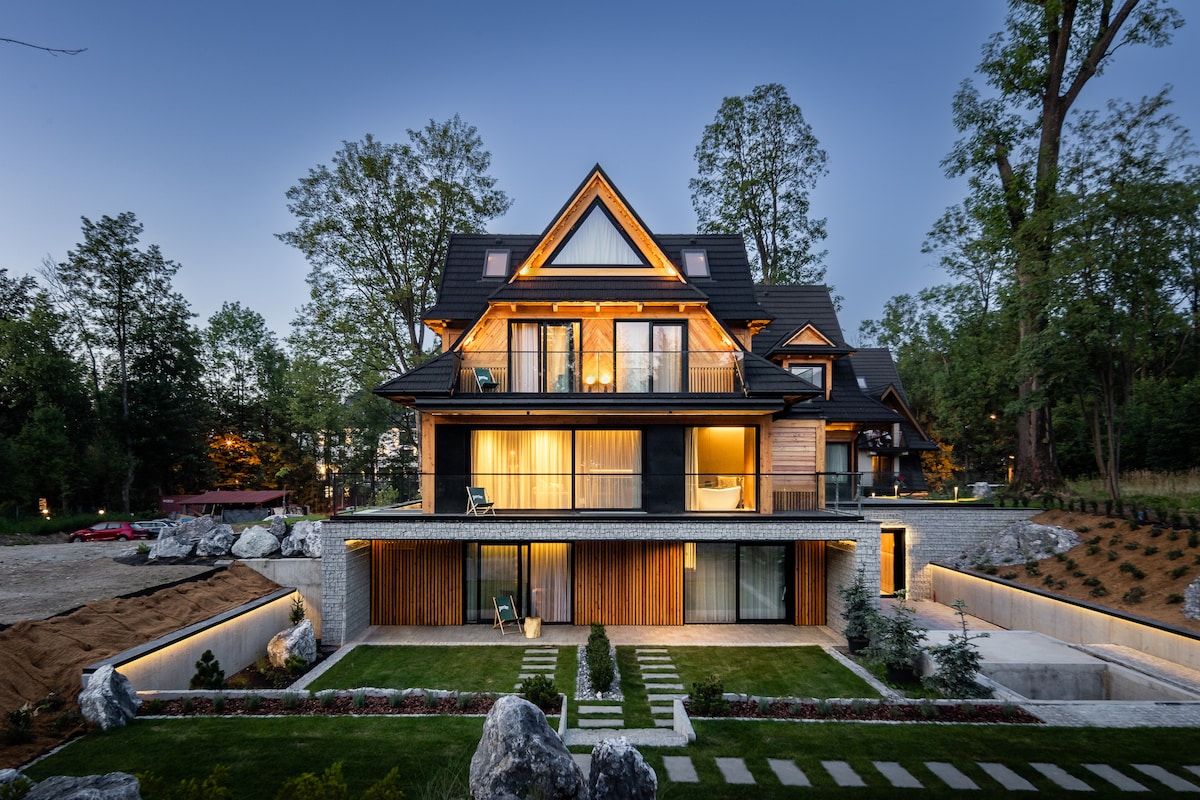 A modern multi-level building is highlighted by exterior lighting during twilight. The façade combines wood and stone elements, with expansive glass windows. The landscaped area features a neatly manicured lawn and stone pathways leading to the entrance, framed by greenery and ornamental trees.