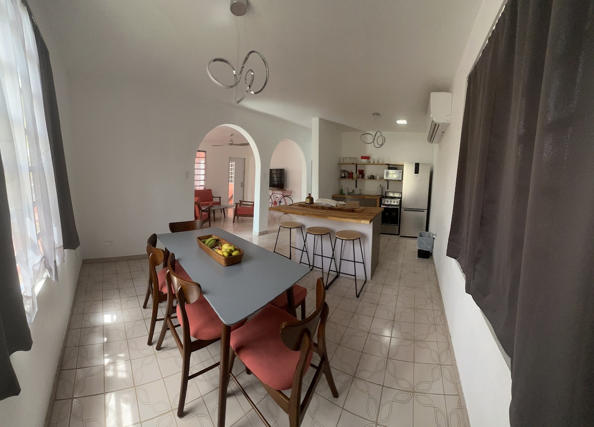 A dining area features a gray table surrounded by wooden chairs with red cushions. Natural light enters through large windows, casting patterns on the tiled floor. An open kitchen can be seen in the background, equipped with modern appliances and a breakfast bar.