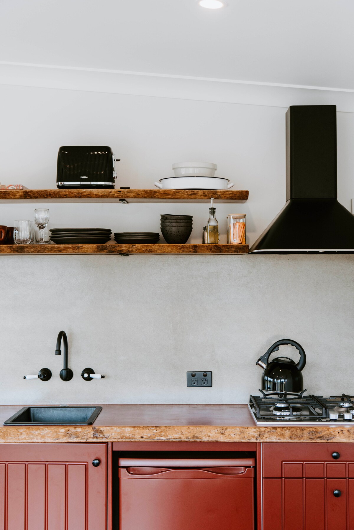 A modern kitchen area is presented with carefully arranged open shelving above vibrant red cabinetry. Kitchen appliances, including a kettle and toaster, are visible alongside an array of plates and glasses. A sleek black sink stands ready beneath stylish wall-mounted taps.