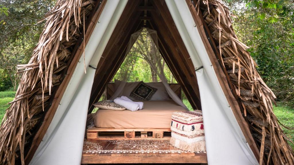A quaint A-frame structure showcases a cozy sleeping area with a spacious bed adorned with neutral linens and decorative pillows. Natural light filters through the open entrance, highlighting earthy wooden elements and surrounding greenery.