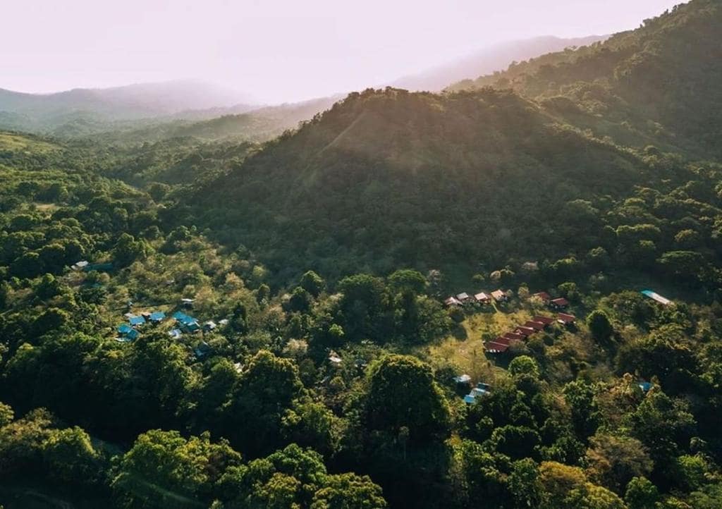 An aerial view showcases a lush, green landscape embraced by rolling hills and mountains. A cluster of cabins with colorful roofs can be seen nestled within a vibrant forest area, indicating a serene and natural retreat amidst diverse foliage.