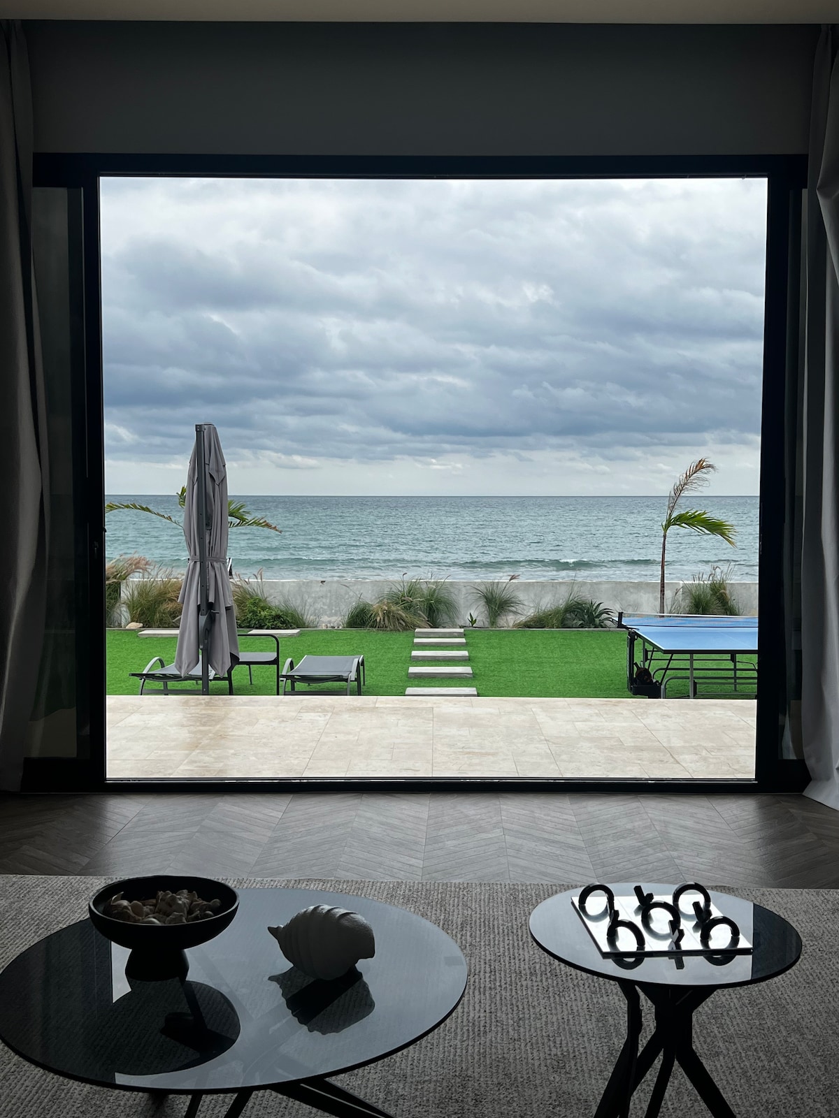 A large glass window frames a serene view of the ocean, with gentle waves lapping at the shore. A well-maintained lawn leads to a wooden deck, where lounge chairs and a sun umbrella are positioned. Cloudy skies add a calm ambiance to the scene.