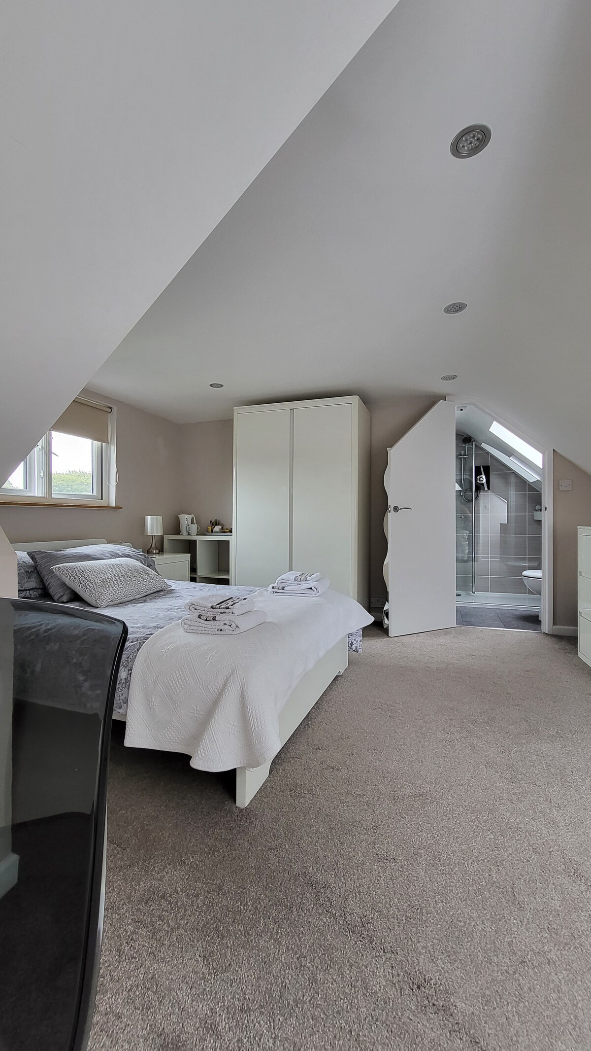 A bright and airy room features a spacious bed with a light blanket, accompanied by folded towels. A wardrobe and nightstand are visible, while a bathroom can be seen through an open door. Natural light enters through a window, enhancing the serene atmosphere.