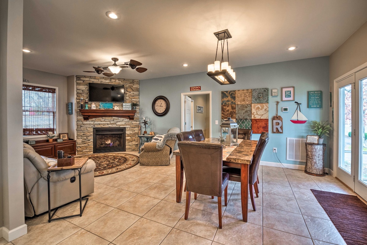 The open living and dining area features a stone fireplace, a large dining table with brown leather chairs, and artistic wall decor. Natural light flows through sliding glass doors that lead to the outdoor space. A comfortable seating arrangement is positioned nearby.