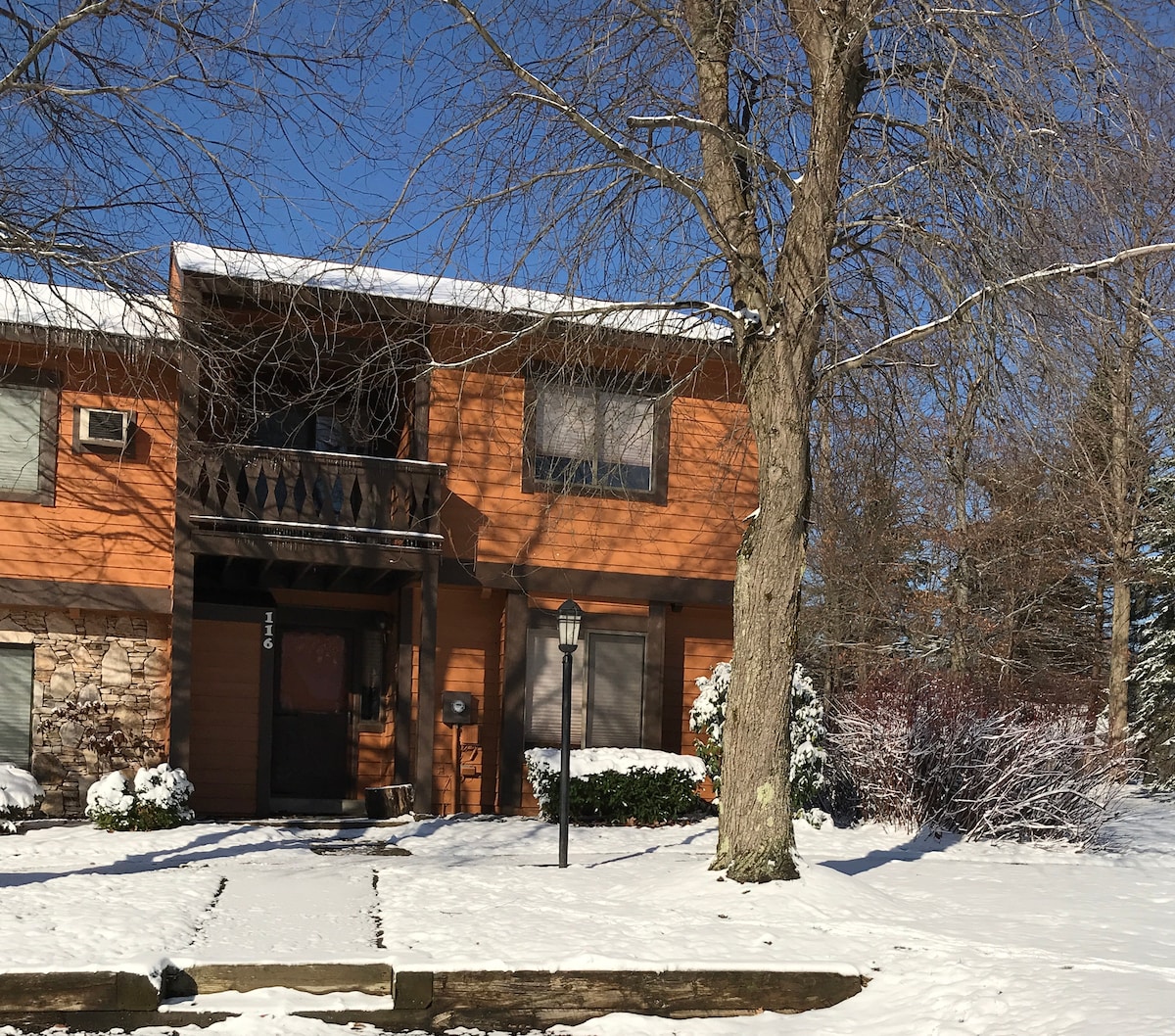 A charming townhome is presented with a snow-covered exterior, highlighting wooden paneling and large windows. The entrance features a dark door framed by a balcony above, with a lamp post nearby. Snow blankets the ground, creating a serene winter atmosphere.