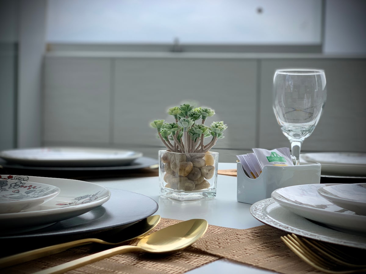 A dining table is set with white and patterned plates, complemented by glassware and gold utensils. A small vase with green plants rests at the center, alongside a white holder containing folded napkins. Natural light enhances the inviting atmosphere.