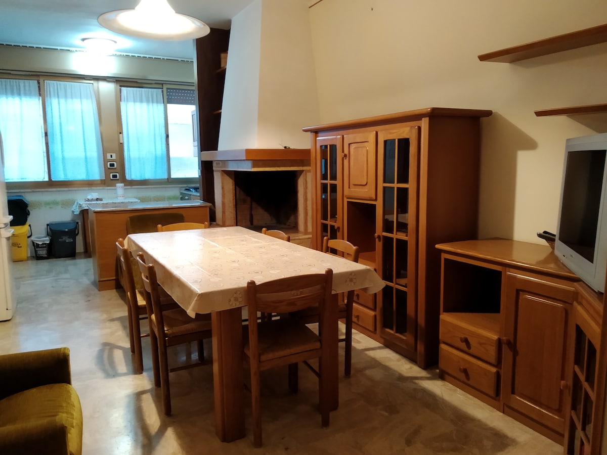 A welcoming dining area features a large wooden table surrounded by chairs. A fireplace is present within a wooden cabinet, complemented by a television unit and additional storage. Natural light enters through window curtains, illuminating the space.