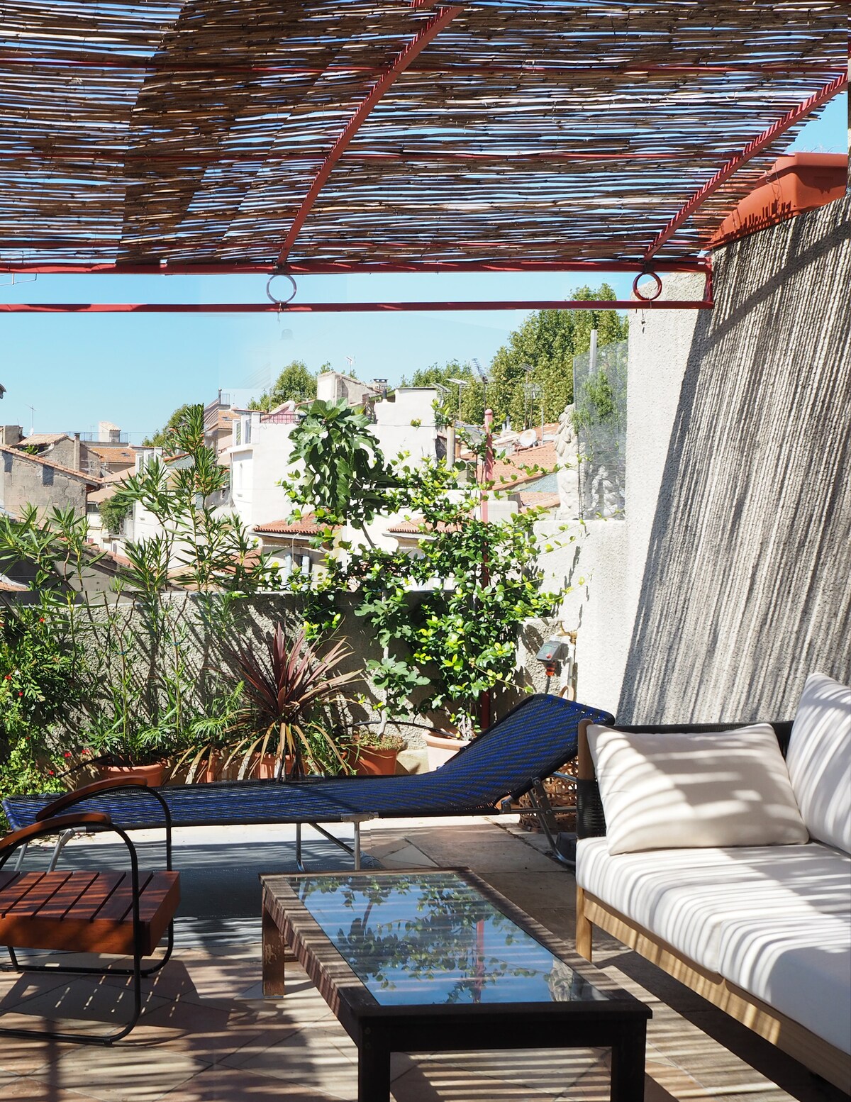 A spacious terrace is visible, featuring a comfortable seating area and a wooden table. Sunlight filters through a woven canopy, casting gentle shadows. Lush plants surround the space, adding greenery, while the view includes rooftops and nearby structures, creating a sense of peaceful outdoor living.