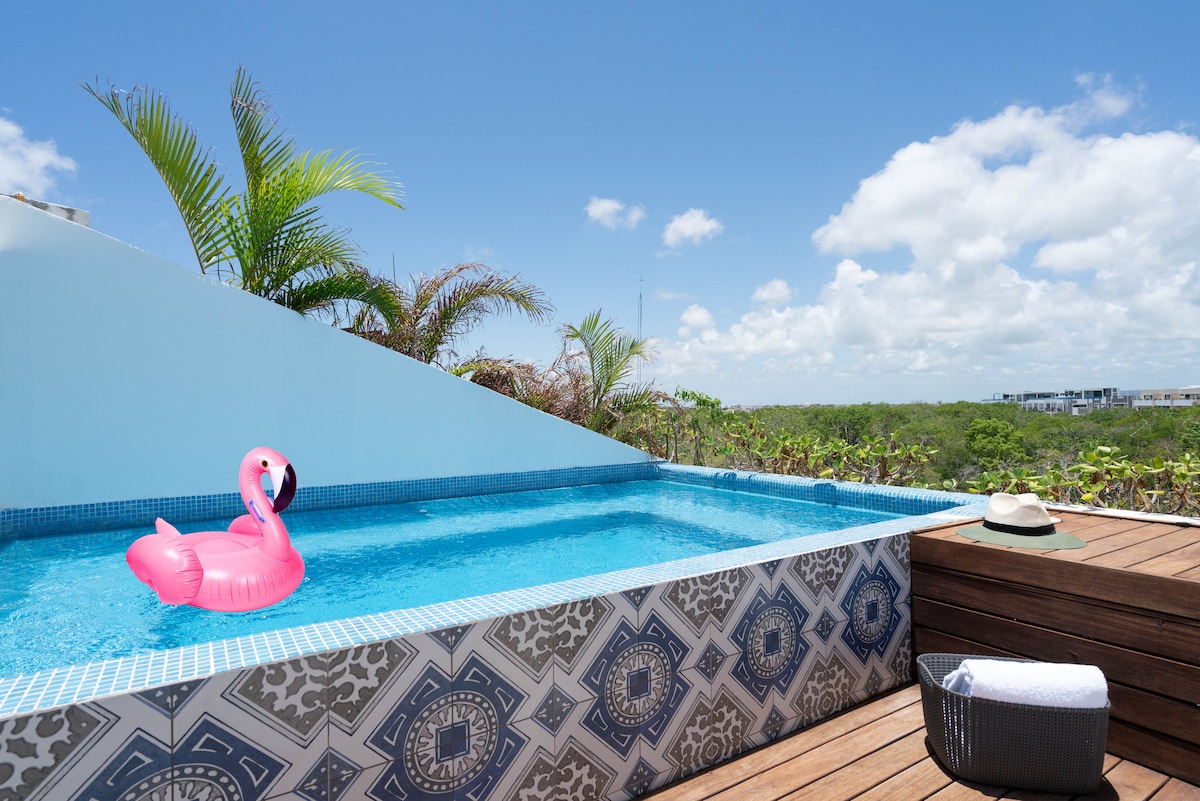 A private pool is situated on a rooftop terrace, adorned with blue tiles and surrounded by greenery. A pink flamingo float is placed in the water, while a sun hat and a rolled towel rest on the wooden deck, reflecting a tranquil outdoor space.