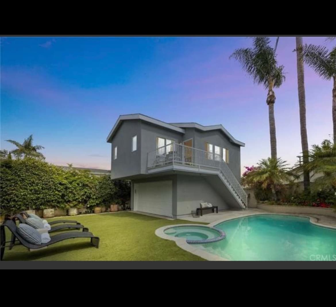A guest house stands elevated with a two-story design, surrounded by lush greenery. A sparkling pool is framed by landscaped grass and palm trees. Lounge chairs offer a space for relaxation, while a staircase leads to the entrance of the upper level.