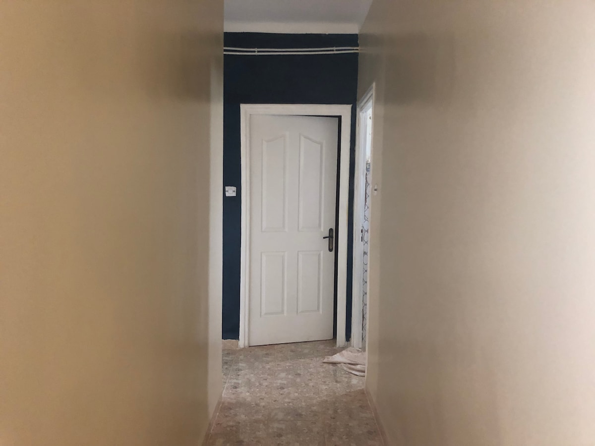 A well-lit hallway features a pale beige wall on the left and a deep navy wall on the right. A white door dominates the end of the corridor, leading into another room. The flooring is covered with textured tiles, adding a subtle depth to the space.