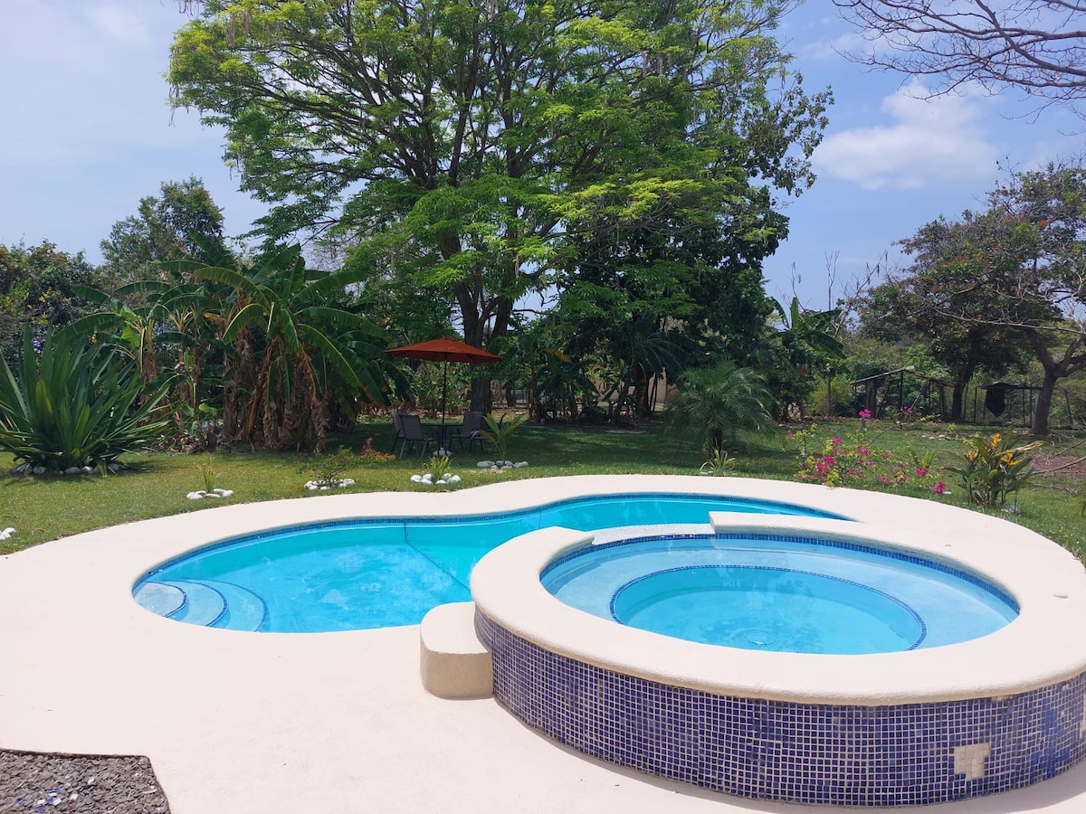 A spacious outdoor area features a two-section swimming pool, surrounded by lush greenery and colorful flowers. A shade structure with an orange canopy is visible in the backdrop, along with various plants and trees providing a tranquil environment.