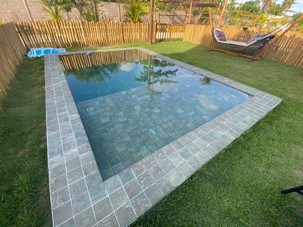 A private swimming pool is framed by a stone-tiled border, set within a lush green lawn. The clear water reflects the surrounding environment. A hammock hangs nearby, accompanied by tropical plants and a wooden fence enhancing the outdoor space.