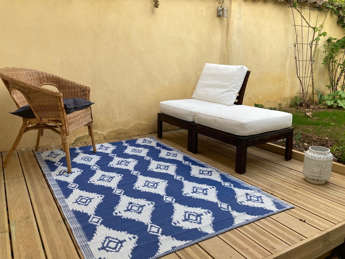 An outdoor terrace area features a wooden deck with a patterned blue rug. A single wicker chair with a cushion is positioned next to a white lounge chair. The surroundings include a soft yellow wall and greenery, providing a calm and inviting outdoor space.