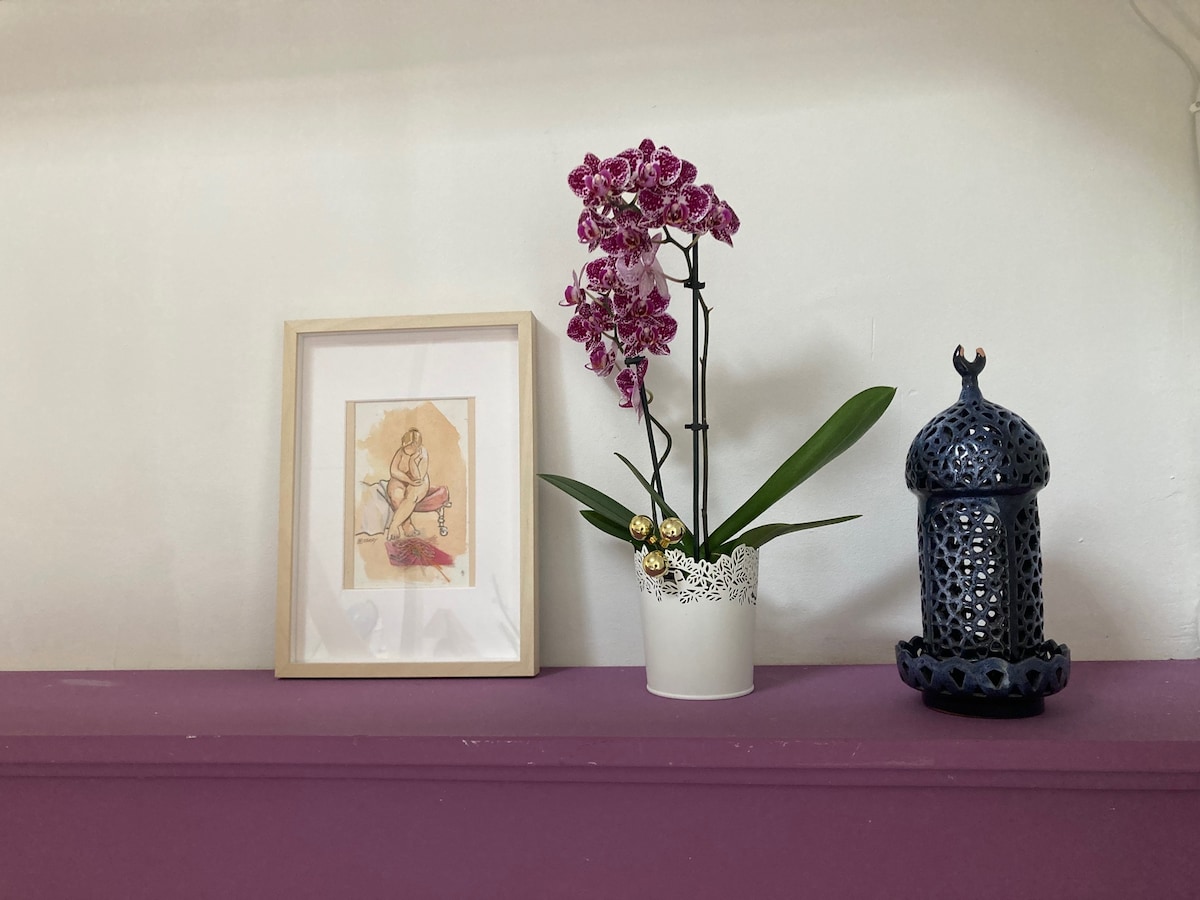 A decorative shelf features a framed artwork alongside a blooming orchid in a simple white pot. A delicate gold bell is attached to the plant. An intricate blue lantern adds an artistic element, enhancing the overall decor with a combination of colors and textures.