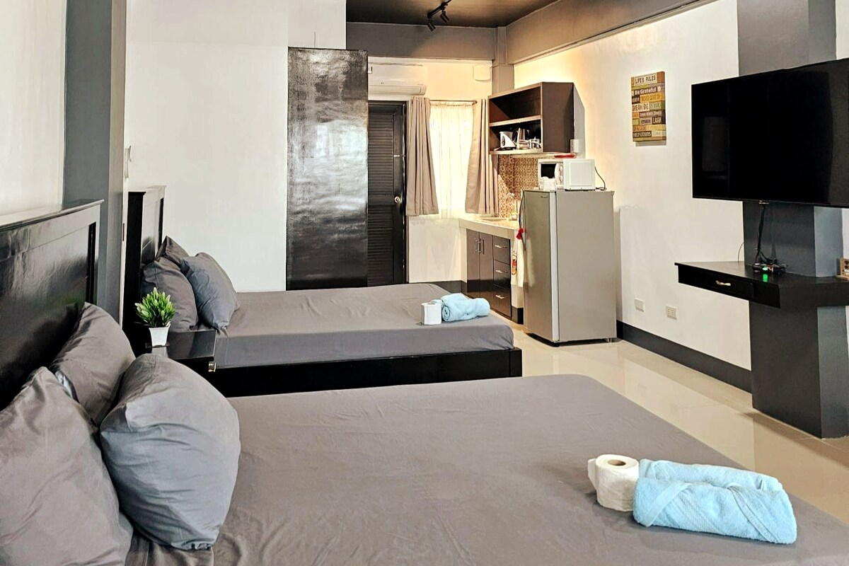 A spacious room features two large beds with gray linens, accompanied by a small plant and rolled towels on a bedside table. A kitchenette with basic appliances is visible in the background, and a wall-mounted TV is positioned for viewing.