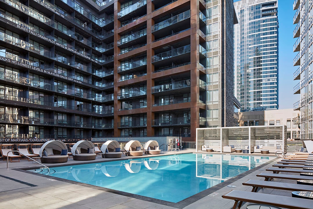 A pristine outdoor pool is surrounded by lounge chairs and contemporary cabanas, featuring modern architecture in the backdrop. Sunlight reflects off the water, highlighting a spacious area designed for relaxation and leisure in an urban environment.
