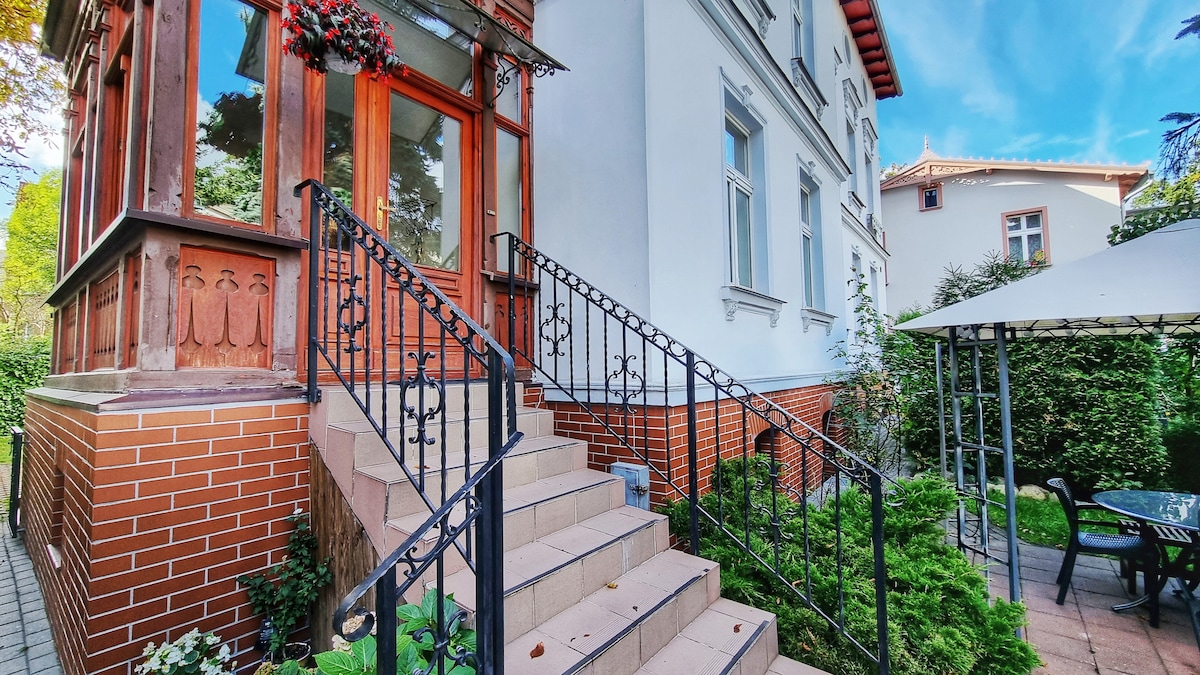 An elegant entrance features a set of wide stone steps leading up to a beautifully detailed façade. The steps are flanked by lush greenery and decorative wrought iron railings. A charming seating area with a canopy is visible in the garden beside the building.