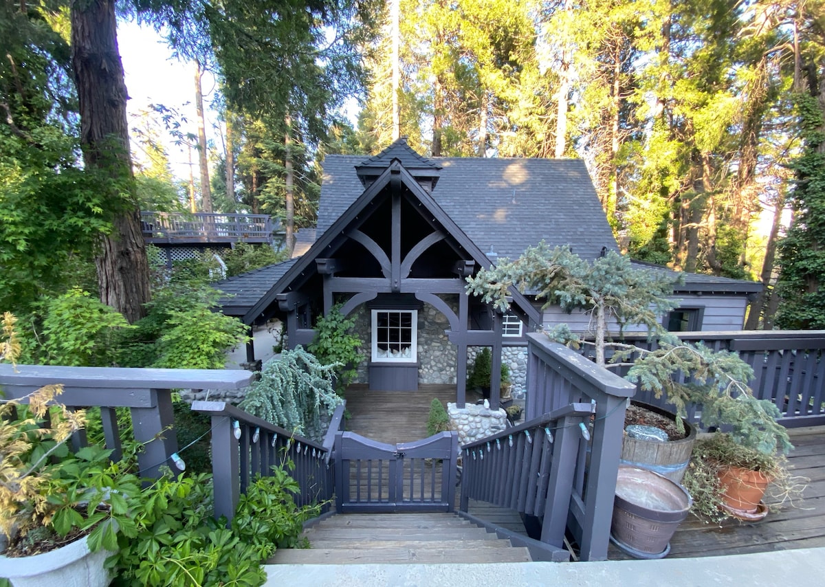 The exterior of the mountain cottage is framed by towering trees and lush greenery. A wooden staircase leads to the entrance, which features a central door surrounded by ornamental plants. The building has a steep roof and exposed timber beams, providing a cozy, rustic charm.