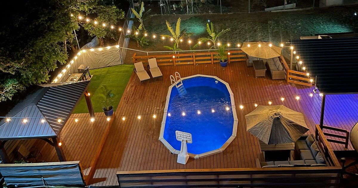 An outdoor area features a vibrant blue pool, surrounded by wooden deck and lounging spaces. Soft lights hang above, creating a cozy atmosphere. Two umbrellas provide shade over seating arrangements, while tropical plants add a touch of greenery to the environment.