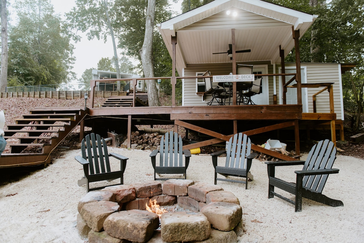 A picturesque outdoor setting features a fire pit surrounded by five Adirondack chairs. In the background, a charming cabin with a covered deck and seating area offers a welcoming retreat. Natural landscaping enhances the tranquility of the scene, with trees providing a serene backdrop.