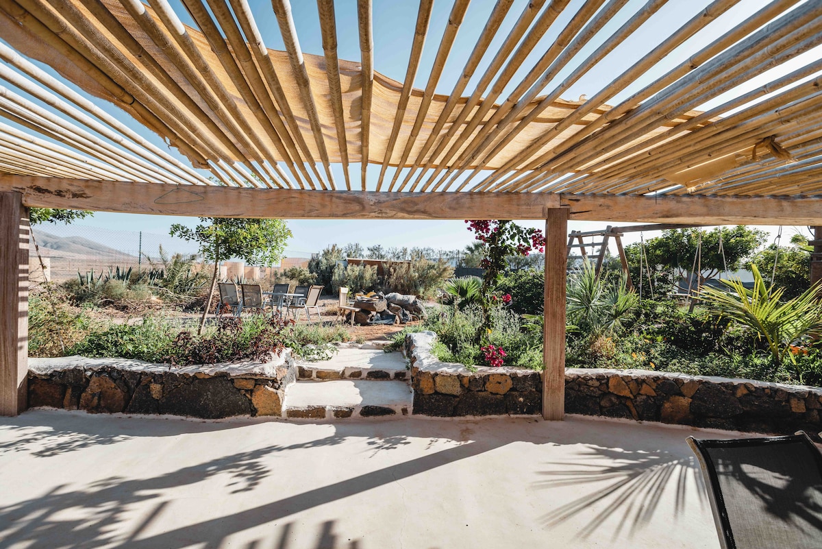 A shaded outdoor area is framed by wooden slats, providing a view of the garden filled with various plants and flowers. Pathways lead through the greenery, and a children's play area with swings is visible in the distance, all under clear blue skies.