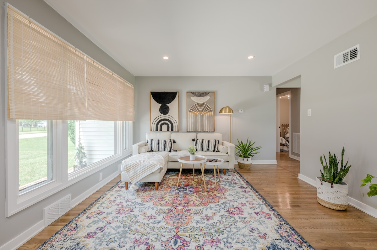 A welcoming living area features a light-colored sofa accompanied by small circular tables. Large windows allow natural light to fill the room, while decorative wall art and indoor plants add interest. A patterned area rug provides a cozy touch to the hardwood floor.